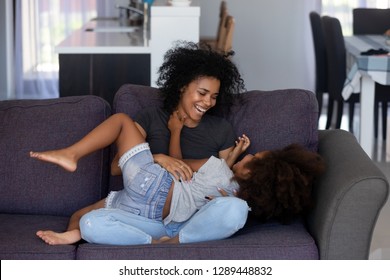 Happy african american mom tickling kid daughter laughing together on couch, cheerful black mother playing funny game with little cute girl at home, mixed race child having fun relaxing with mum