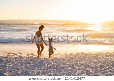 Happy african american mother with cute little girl at beach walking towards the sea. Black family walking towards ocean on beach during sunset with copy space. Mother and child running barefoot.