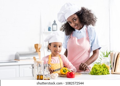 happy african american mother and daughter in chef hats cutting bell pepper on kitchen