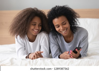 Happy African American Mother And Daughter Lying On Bed Looking At Smartphone, Watching Funny Video, Excited Mom And Teen Girl Relax At Home, Using Cellphone, Having Fun Laughing In Bedroom