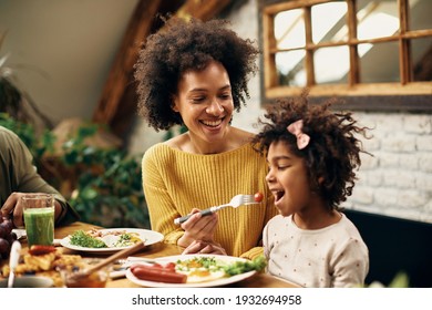 Happy African American mother enjoying while feeding daughter during breakfast in dining room.  - Powered by Shutterstock