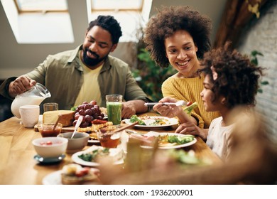Happy African American mother feeding daughter during family meal at dining table. 