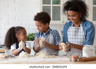 Happy African American mother with little kids wearing aprons kneading dough, cooking baking at home, standing at table in kitchen, smiling mom with 5s daughter and son preparing cookies together - Powered by Shutterstock