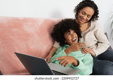 Happy African American Mother Looking At Excited Daughter Lying With Laptop On Couch