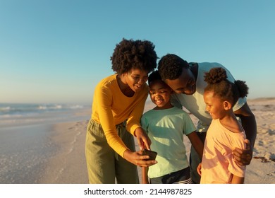 Happy african american mother sharing smartphone with family at beach on sunny day. unaltered, family, lifestyle, togetherness, technology and holiday concept.