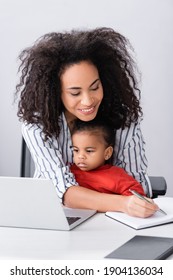 happy african american mother sitting with toddler daughter while writing in notebook neat laptop on desk