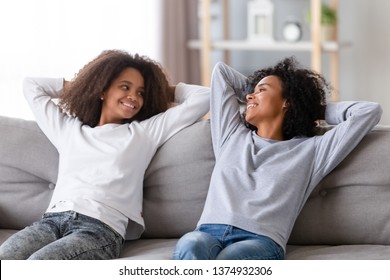 Happy African American Mother And Teen Daughter Relaxing Together At Home, Leaning Back With Hands Behind Head On Cozy Sofa, Smiling Black Mum And Teenage Girl Talking, Family Weekend