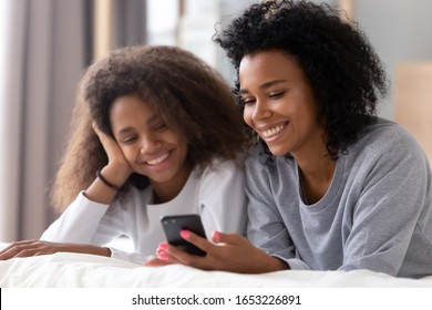 Happy African American Mother And Teen Daughter Using Smartphone Apps For Shopping Social Media Lying On Bed, Smiling Black Mom With Teenage Girl Having Fun Watch Videos On Phone Laughing In Bedroom