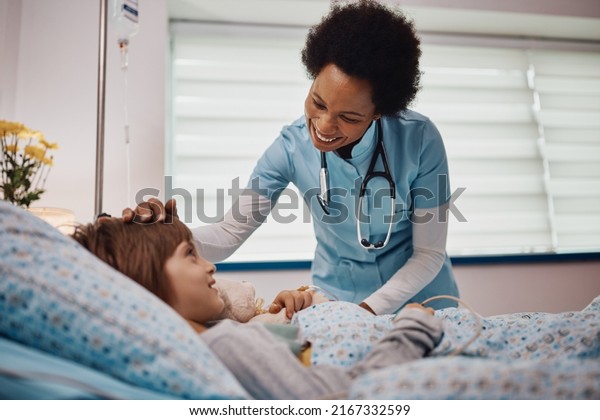 Happy African American nurse\
communicating with a child who is recovering at hospital\
ward.