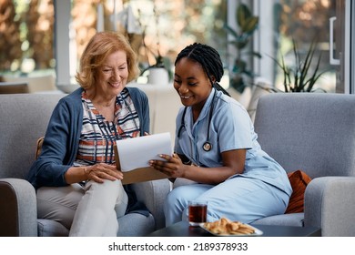 Happy African American Nurse And Senior Woman Going Through Medical Insurance Paperwork At Nursing Home.