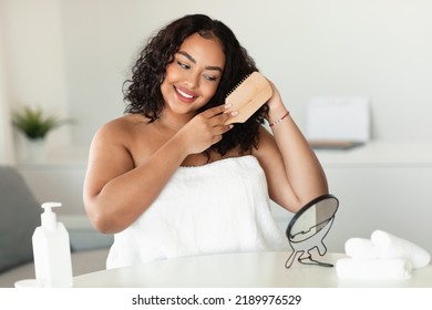 Happy African American Oversize Woman Brushing Her Curly Hair With Comb While Sitting Near Mirror In Bedroom. Positive Black Lady Wrapped In Towel Making Haircare Routine