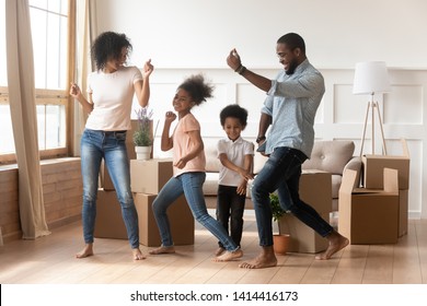 Happy african american parents and cute children dancing among boxes celebrating moving day relocation renovation, active carefree funny mixed race family mom dad having fun with kids in new house