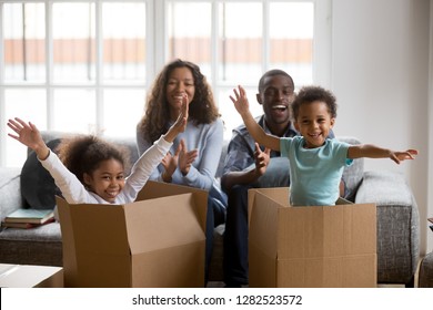 Happy African American Parents And Kids Playing In Boxes Enjoy Relocation Into New Home, Excited Mixed Race Children Having Fun Help Mom Dad Unpack In Living Room, Black Family On Moving Day Portrait