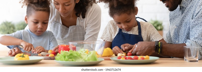 Happy African American parents teaching sibling kids to cook, slicing fresh vegetables, peppers, tomatoes for salad on kitchen table or counter. Family healthy eating, organic food concept. Banner - Powered by Shutterstock