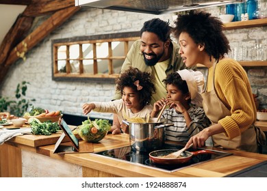 Happy African American parents and their kids having fun while cooking and following recipe on the internet. 