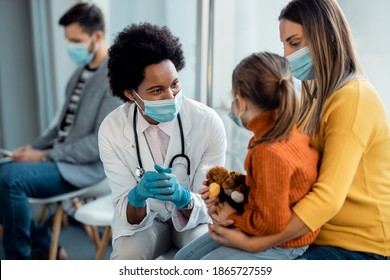 Happy African American pediatrician with face mask talking to mother and daughter in a waiting room at the hospital.