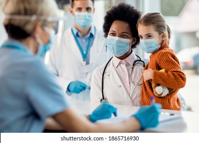 Happy African American Pediatrician And Little Girl Communicating With Nurse At Reception Desk In The Hospital While Wearing Protective Face Masks.