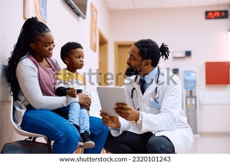 Happy African American pediatrician using digital tablet while communicating with a boy and his mother in hallway at medical clinic. 