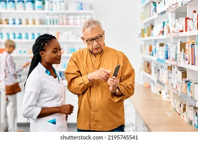 Happy African American Pharmacist Assisting Senior Patient Who Is Using Mobile App On Smart Phone In Drugstore. 