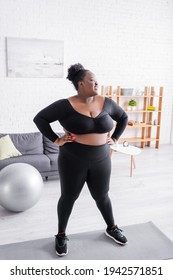 Happy African American Plus Size Woman In Sportswear Standing With Hands On Hips In Living Room