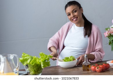 happy african american pregnant woman making salad at kitchen