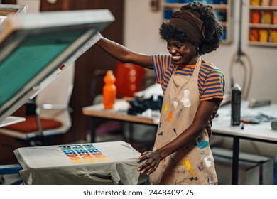 Happy african american professional graphic technician is finishing silk screen printing on t-shirt at printing shop. Smiling multicultural graphic technology expert is smiling at finished product. - Powered by Shutterstock