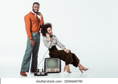 happy african american retro styled girlfriend sitting on vintage television and and talking by phone on white