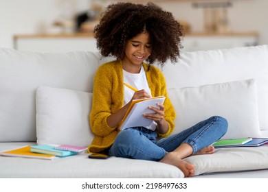 Happy African American School Girl With Bushy Hair Sitting On Couch, Holding Notepad And Pen, Writing Essay And Smiling, Kid Doing Homework, Enjoying Educational Process, Home Interior, Copy Space