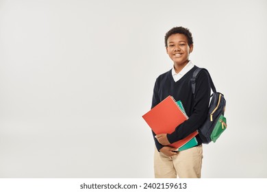 happy african american schoolboy in uniform holding notebooks and standing on grey background - Powered by Shutterstock