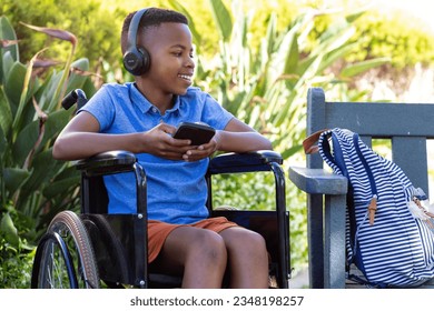 Happy african american schoolboy in wheelchair using headphones and smartphone in garden, copy space. Education, childhood, inclusivity, elementary school and learning concept. - Powered by Shutterstock