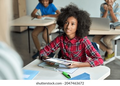 Happy African American schoolgirl looking listening to teacher using tablet device sitting in classroom with group of schoolchildren using laptop computers. Modern technologies for education concept. - Powered by Shutterstock