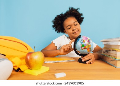 Happy african american schoolgirl sitting at desk in classroom and looking at world map globe on blue background. Back to school concept. - Powered by Shutterstock