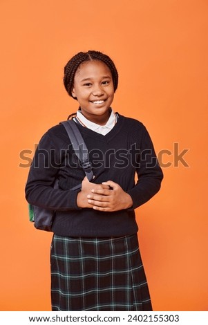 happy african american schoolgirl in uniform smiling and holding backpack on orange background