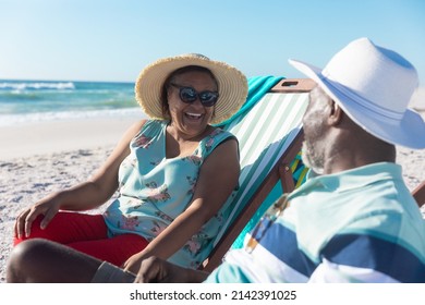 Happy african american senior couple talking while sitting on folding chairs at beach. unaltered, love, togetherness, lifestyle, enjoyment and holiday concept.