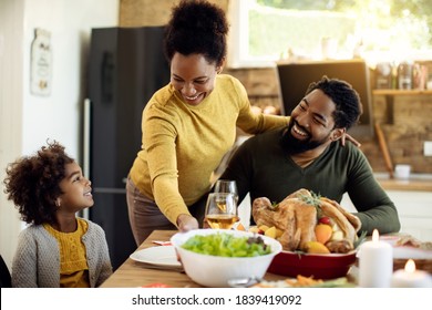 Happy African American Serving Salad To Her Family During Thanksgiving Lunch At Dining Table.