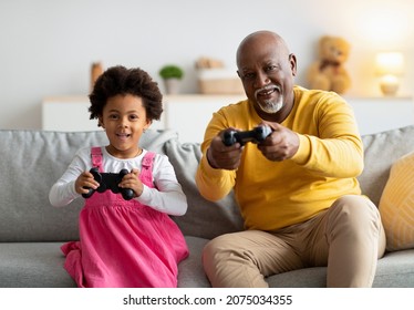 Happy African American Small Girl And Old Man With Joysticks Play In Online Games In Living Room Interior. Entertainment At Home, Have Fun In Spare Time, Copy Space. Grandpa And Granddaughter Together