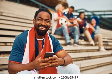 Happy African American Sports Fan Texting On Mobile Phone Outdoors And Looking At Camera.