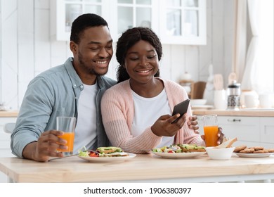 Happy African American Spouses Using Smartphone While Having Breakfast In Kitchen, Cheerful Black Couple Enjoying Tasty Meal And Browsing Internet Or Shopping Online With Cellphone, Free Space - Powered by Shutterstock