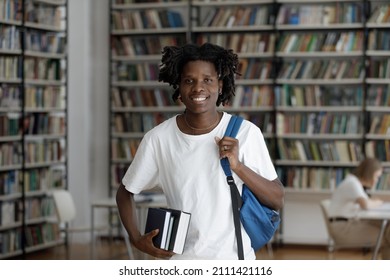 Happy African American Student Guy With Dreads And Backpack Standing In College Library With Bookshelves Behind, Holding Stacked Books, Looking At Camera, Smiling, Head Shot Portrait