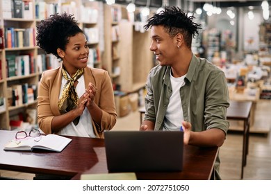 Happy African American student using laptop and talking to his professor who is congratulating him on well done assignment in a library.