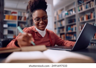 Happy African American student using laptop and taking notes while studying in library. - Powered by Shutterstock