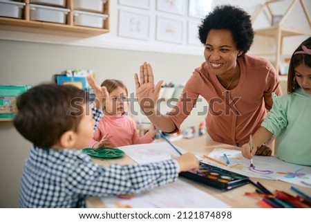 Happy African American teacher and small boy giving high-five during art class at kindergarten. 