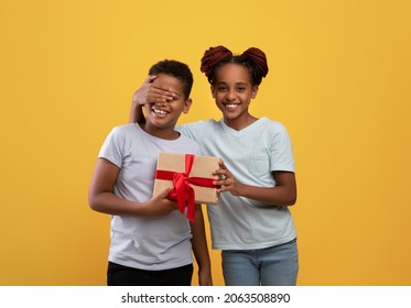 Happy African American Teen Girl Loving Sister Greeting Her Brother With Birthday, Closing Boy Eyes And Giving Gift Box, Yellow Studio Background. Black Siblings Celebrating Birthday Together