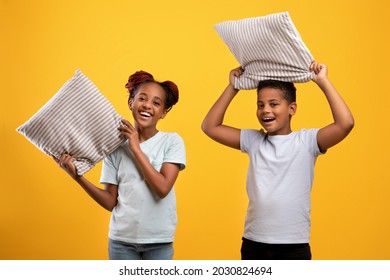 Happy African American Teen Sister And Brother Holding Pillows Over Yellow Studio Background, Cheerful Black Siblings Or Twins Having Pillow Fight, Enjoying Time Together. Siblings Relationships