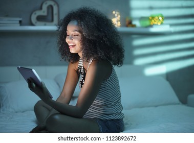 Happy african american teenager in bed at home with tablet. Young black woman watching movie in bedroom at night and laughing.