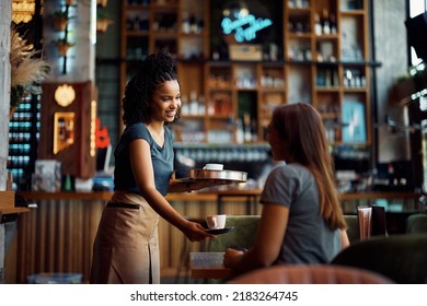 Happy African American waitress serving coffee to female customer while working in a cafe. - Powered by Shutterstock