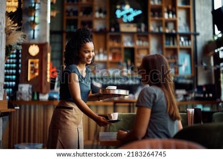 Happy African American waitress serving coffee to female customer while working in a cafe.