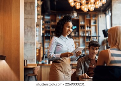 Happy African American waitress using touchpad while taking order from customers in a restaurant. - Powered by Shutterstock