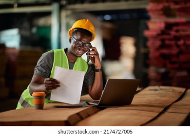 Happy African American warehouse worker going through paperwork while communicating on cell phone and using laptop t wood compartment. 