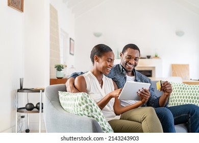 Happy African American Wife Doing Shopping Online While Using Digital Tablet And Credit Card At Home With Husband. Middle Aged Black Couple Making An Online Purchase Using Debit Card On Digital Tablet